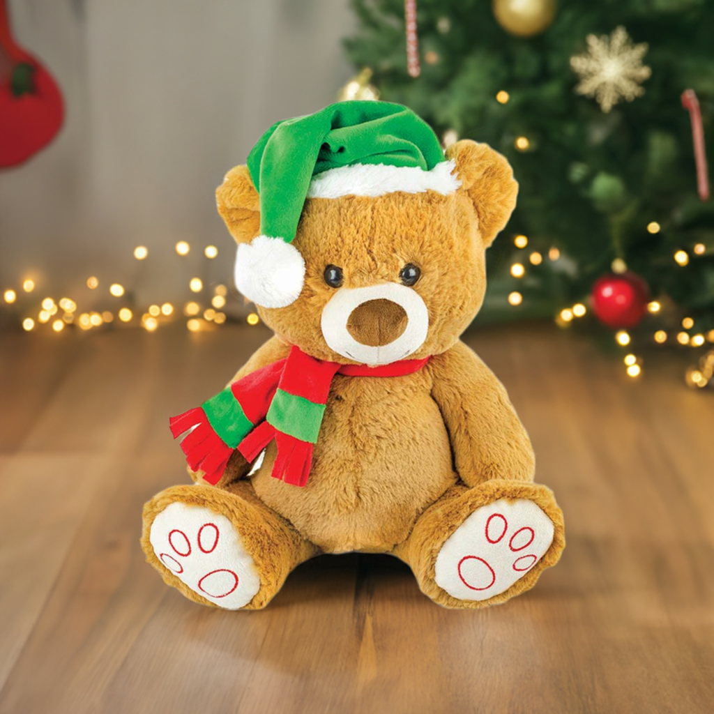 Adorable Prestige Christmas Bear Plush Dog Toy featuring a green hat, and red scarf, sitting on a wooden floor next to a Christmas tree.