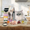 An adult cat on a wooden tabletop next to a bag of Black Hawk Hairball food, featuring raw chicken, rice, egg, liver, and oils in the foreground.