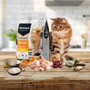 A healthy adult cat on a wooden tabletop next to a bag of Black Hawk Weight Management food, featuring fresh chicken and high-fibre ingredients displayed in front.