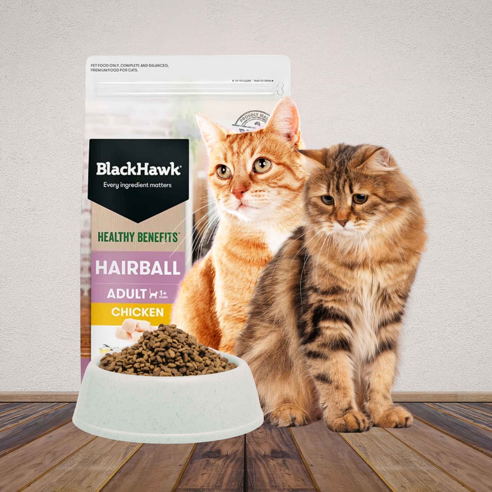 Two cats sitting on a wooden floor behind a bowl of dry cat food with a bag of Black Hawk Healthy Benefits in the background.