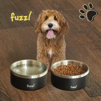Small dog sitting on a wooden floor between two black and silver Fuzzl Titan Thermal Dog bowls with 'Fuzzl' branding. 