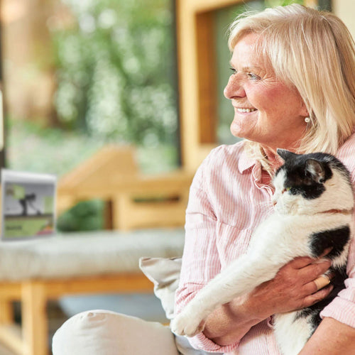 Lady holding a cat and smiling - a blurred bag of Meals for Meows Single Protein Duck Cat Food is in the background.