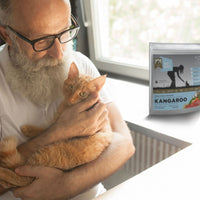 Man holding a cat next to a package of Meals for Meows Kangaroo Cat Food 2.5kg.