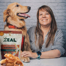 A happy dog sitting with their owner next to a pack of Zeal Air-Dried Dog Treats - Chewies, with loose treats placed in front of the pack. Highlighting the premium, free-range New Zealand-made product.