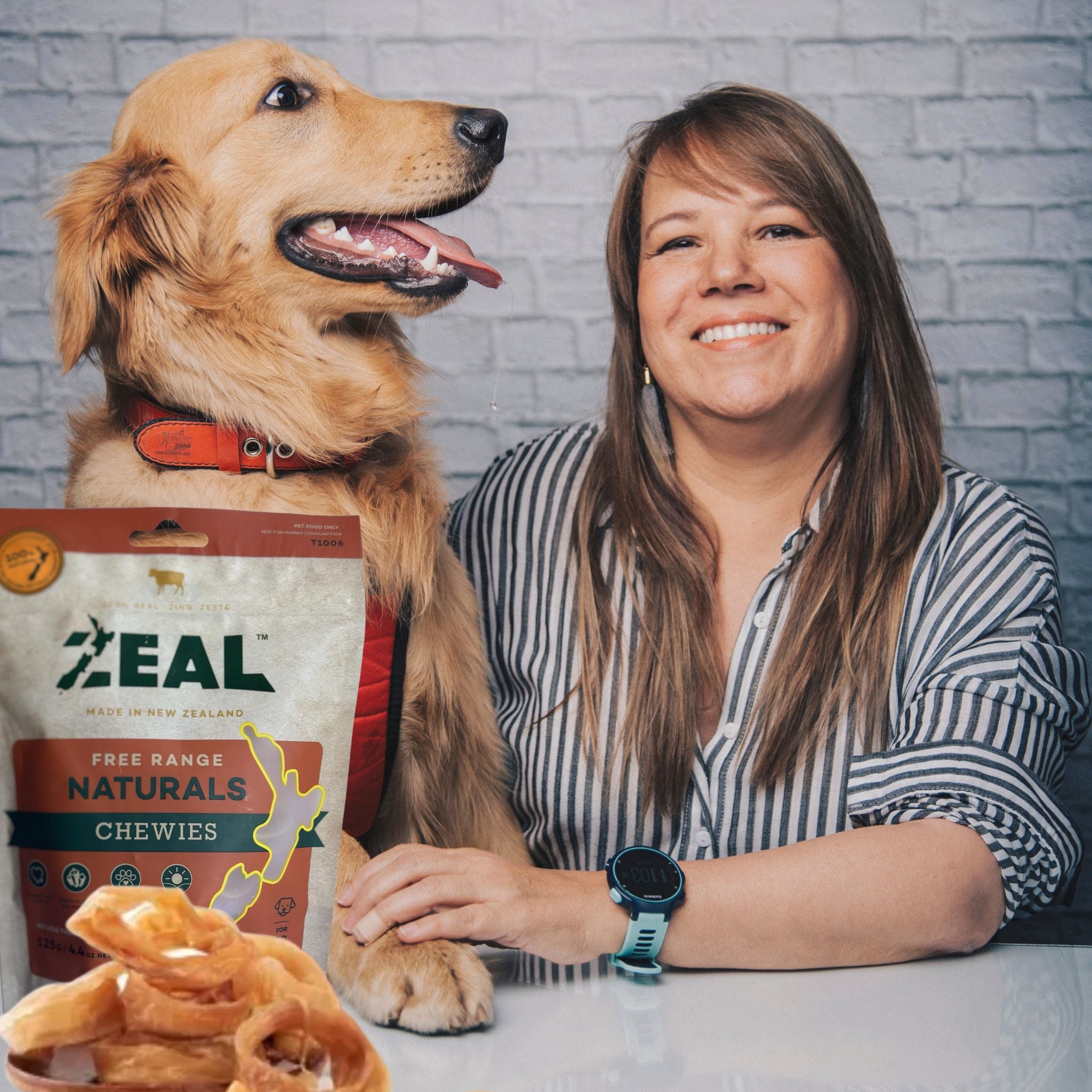 A happy dog sitting with their owner next to a pack of Zeal Air-Dried Dog Treats - Chewies, with loose treats placed in front of the pack. Highlighting the premium, free-range New Zealand-made product.