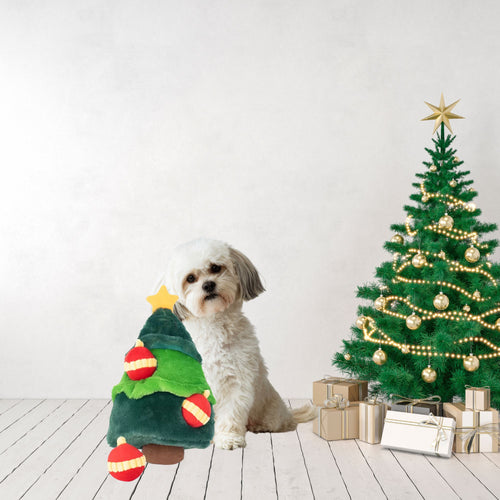 Small white dog sitting beside the ZippyPaws Holiday Burrow Christmas Tree dog toy in a festive room with a decorated Christmas tree and gifts