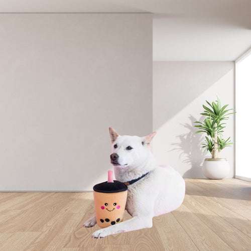 A white dog lies on a light wood floor in a bright room, holding a ZippyPaws NomNomz Boba Milk Tea Plush Dog Toy with a black lid and pink straw. The cup has cute embroidered facial features.