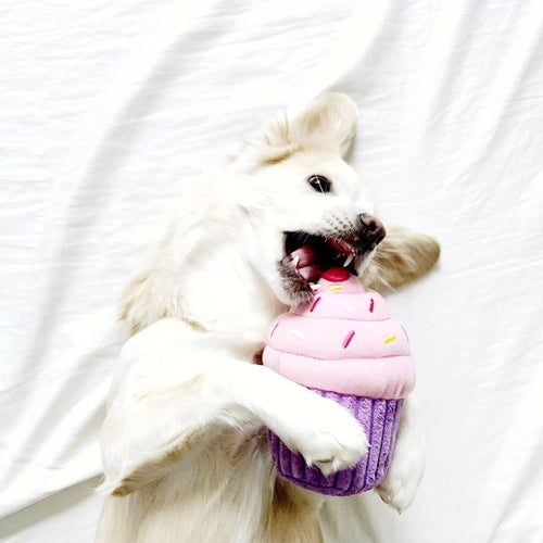 A dog biting into the pink NomNomz Cupcake plush toy with a purple base while lying on a white bedsheet.