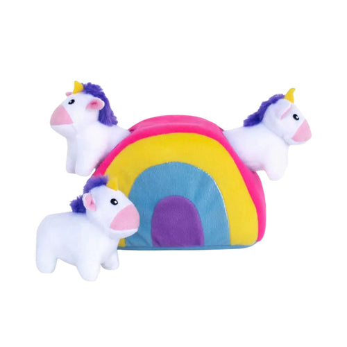 A colourful plush rainbow-shaped burrow dog toy by ZippyPaws. Two small white plush unicorn toys poke their heads out of holes in the rainbow structure, while a third plush unicorn stands upright on the surface directly in front of the burrow.